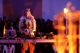 Fotografie eines Mannes in Kostüm an einem DJ Pult in lila orangener Beleuchtung.