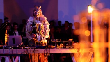 Fotografie eines Mannes in Kostüm an einem DJ Pult in lila orangener Beleuchtung.