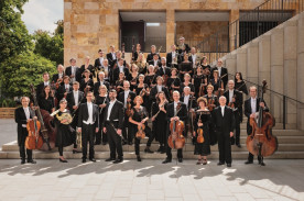 Mehrere Musiker*innen stehen schwarz/weiß gekleidet auf einer Treppe. Einige Musiker*innen halten Instrumente in der Hand.
