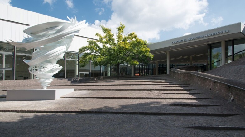 Eingangsbereich des Sprengel Museums Hannover. Auf der linken Seite ist ein weißes Kunstwerk und ein Baum zu sehen. Der Himmel ist blau mit ein paar Wolken und die Sonne scheint.