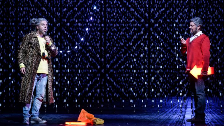 Zwei Männer stehen gegenüber auf der Bühne und sehen sich an. Einer hat eine Flöte in der Hand. Der Hintergrund ist mit Lichterketten behangen.