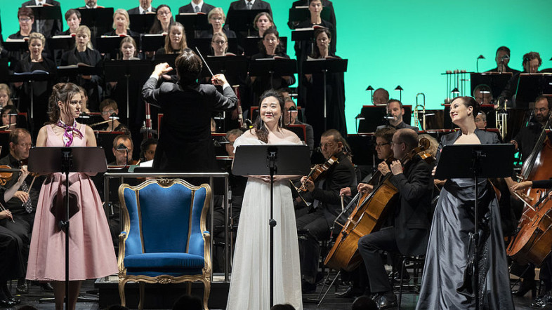 Im Vordergrund stehen zwei Frauen jeweils vor einem Podest. Im Hintergrund ist ein Orchester zu sehen.
