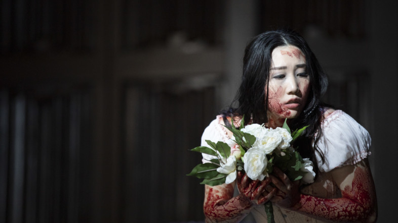 Eine asiatische Frau mit einem, blutverschmierten weißen Kleid hält weißen Blumen in ihrer Hand und wirkt verängstigt.