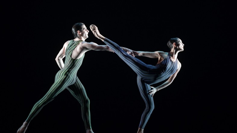 Zwei Ballett-Tänzer tanzen in Kostümen auf einer Bühne vor einem schwarzem Hintergrund.