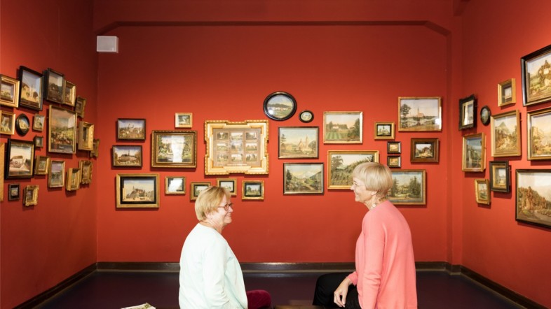 Zwei Frauen sitzen vor einer roten Wand, an der mehrere Bilder hängen. Sie schauen sich an und unterhalten sich.