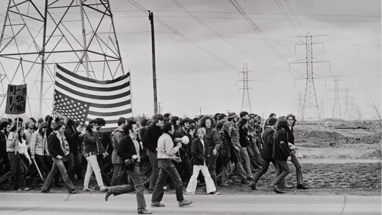 Einen schwarz-weiß Fotografie, auf der eine Gruppe von Menschen auf der Straße läuft. Im Hintergrund sind Strommaste zu sehen.