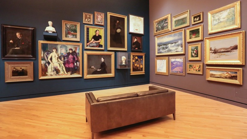 Foto eines Ausstellungsraumes, Im Raum steht eine braune Bank. Eine Wand ist mit blauer Farbe gestrichen, die andere mit einer hellbraunen Farbe. An den Wänden hängen mehrere Bilder.