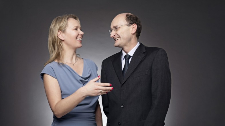 Eine Frau in einem blauen Kleid und ein Mann in einem schwarzen Anzug stehen vor einer dunklen Leinwand. Die beiden schauen sich an und lachen.