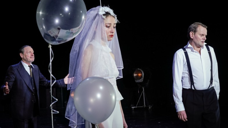 Auf einer Bühne steht eine Frau im Brautkleid, die weint. Links neben ihr sind durchsichtige Ballons. Der Hintergrund ist schwarz und zwei Männer sind zu sehen.