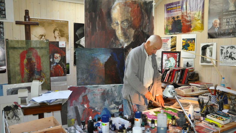 Ein älterer Mann steht in einem Raum voller Gemälde.