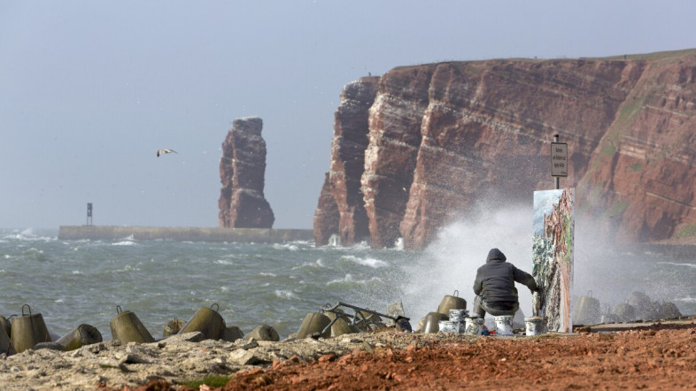 Ein Mann sitzt am Meer und malt auf einer Leinwand. Im Hintergrund sind große Felsen zu sehen.