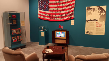 Bild eines nachgebauten Wohnzimmers, an der Wand eine große amerikanische Flagge auf die jemand "Hände weg von Mittelamerika" geschrieben hat. Reil der Ausstellung Auf den Spuren des Kalten Krieges im Freilichtmuseum Molfsee.